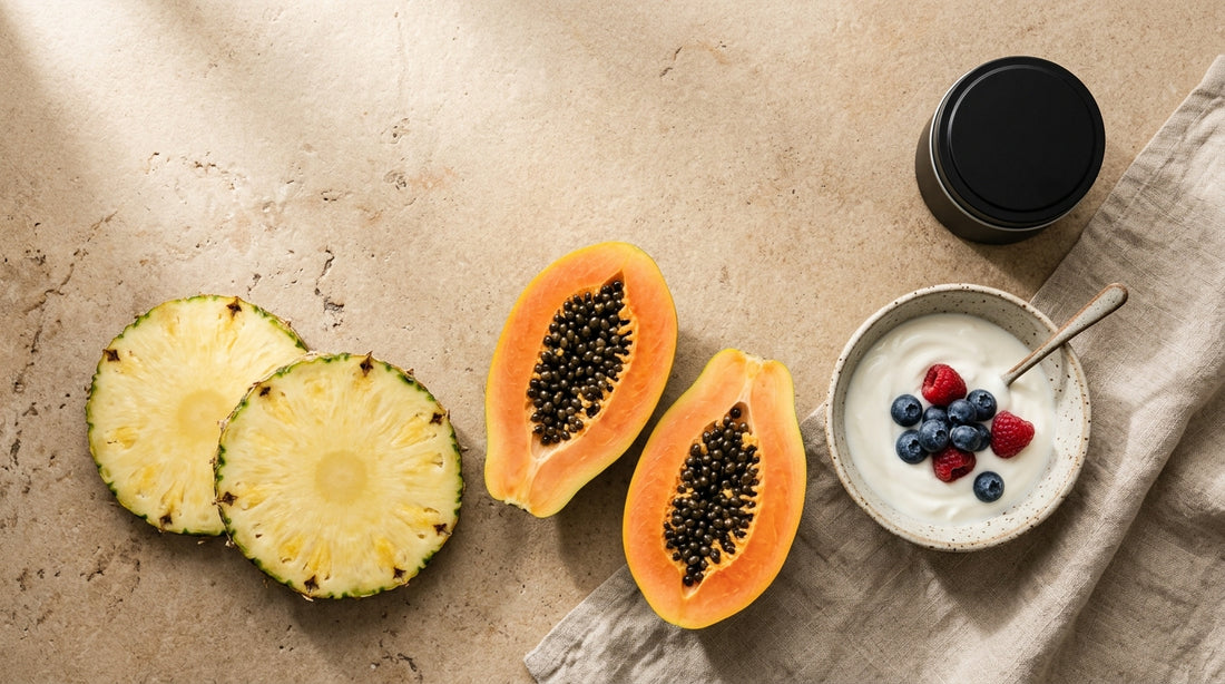 Fresh pineapple rings, halved papaya with seeds, a ceramic bowl of yogurt with berries, and a small matte black supplement tin on a warm stone surface